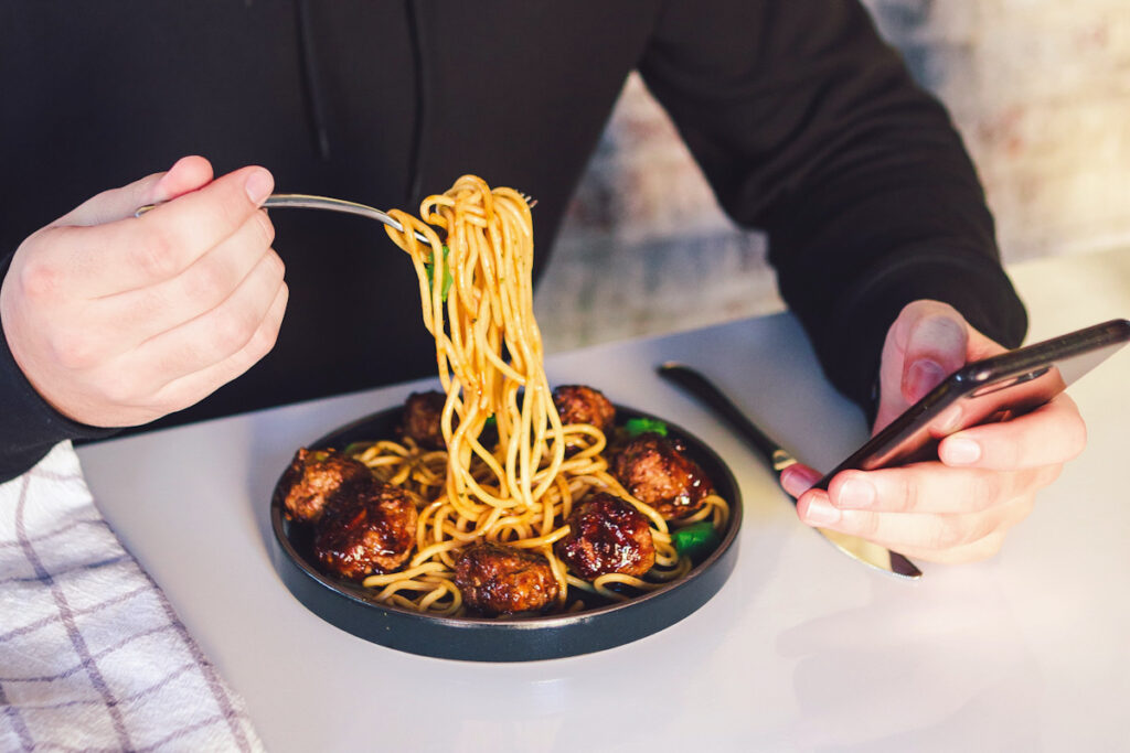 Photo shows someone looking at their phone and eating meatballs with spaghetti at the dinner table. Eating more vegan food (or adopting a flexitarian diet) comes with several benefits.