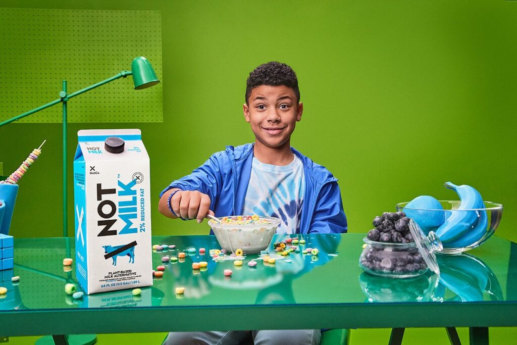 Photo shows a child wearing a blue jacket eating a bowl of cereal. A carton of NotMilk sits on the table next to the bowl.