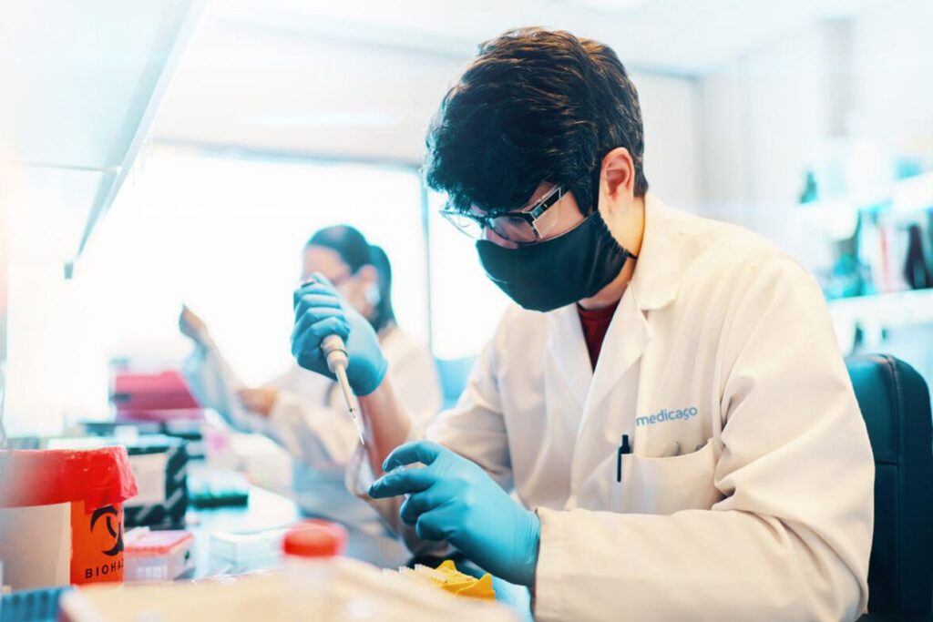 Photo shows a scientist in blue gloves and a black mask working with a syringe.