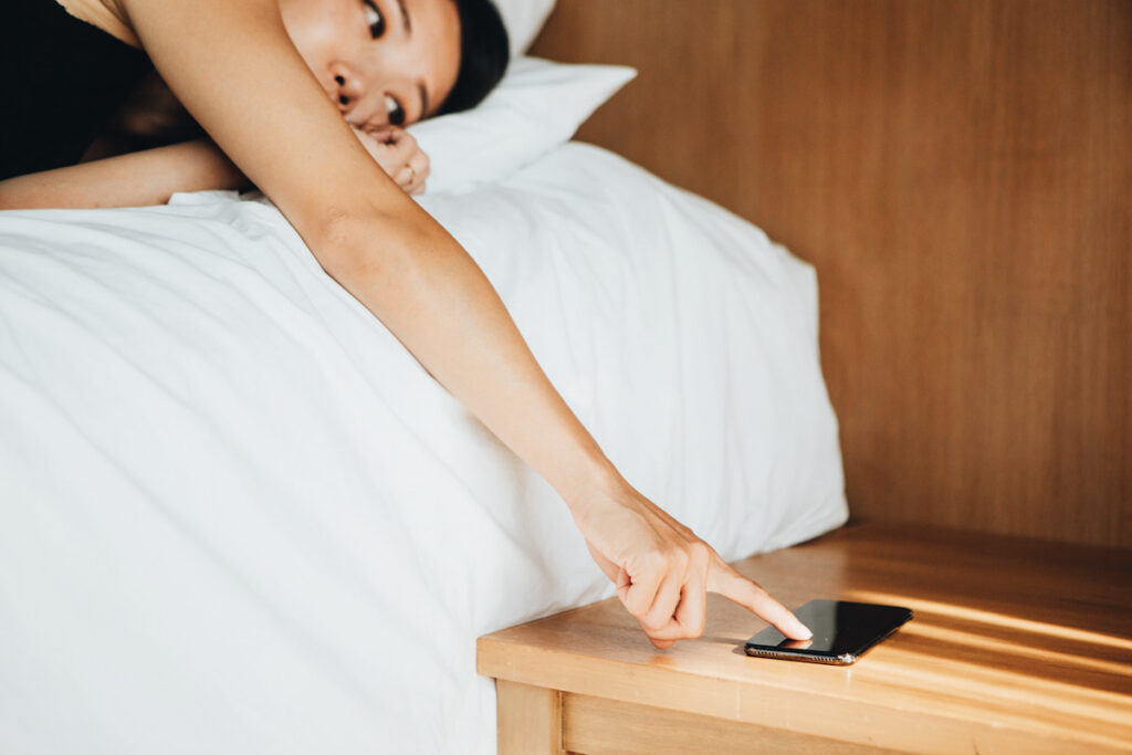 Photo shows a young Asian woman turning off the alarm on her mobile phone while waking up in bed in the morning. Self care can look different for everyone.