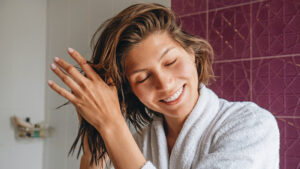 Photo shows a woman using hair products in the bathroom. Using no rinse conditioner can help save on water.