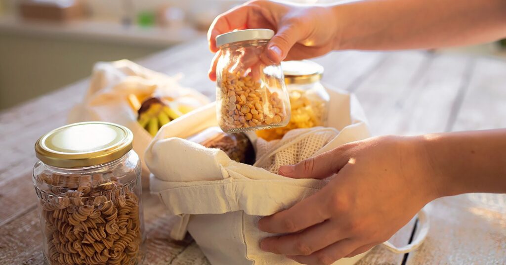 Photo shows a reusable tote filled with mason jars that contain dry pantry goods like split yellow peas and pasta.